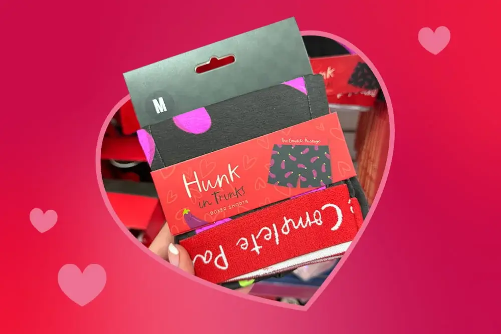 Valentine‑themed men’s boxer shorts in playful heart patterns, held up in front of a store display with a pink, heart‑filled background.