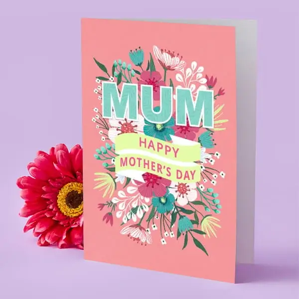 A coral Mother’s Day card decorated with colourful flowers around the word “Mum,” displayed upright beside a bright pink flower.
