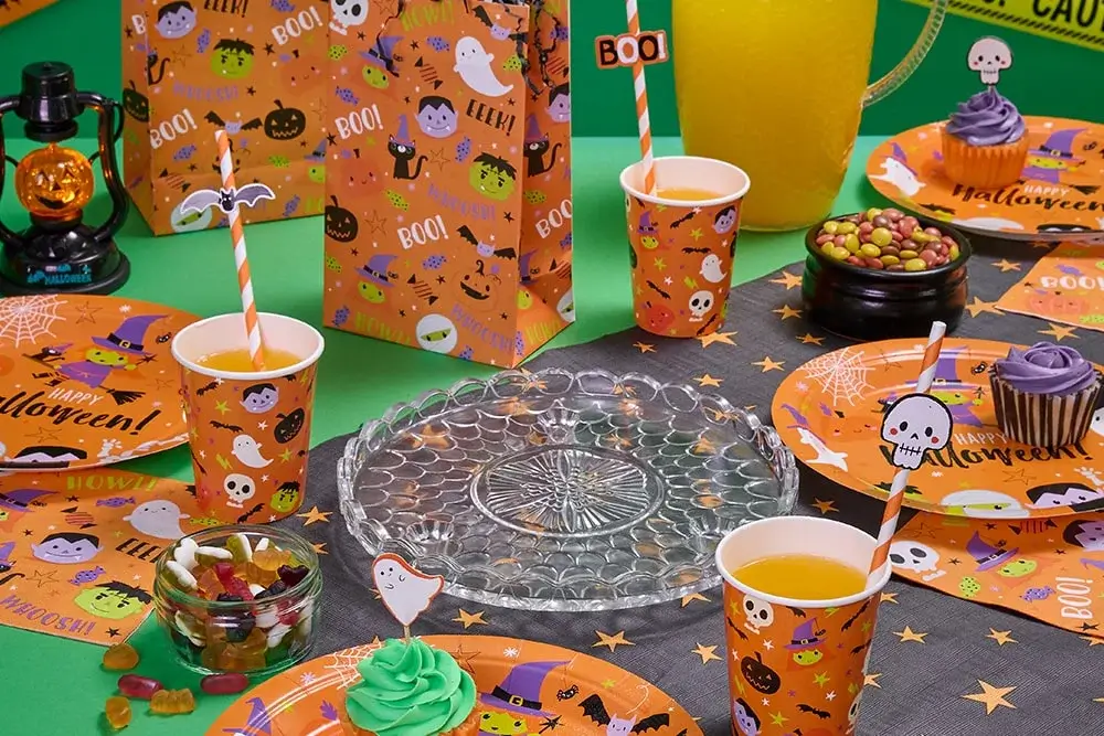 Halloween party table with orange plates, cups, and napkins featuring ghosts, bats, pumpkins, and the word BOO, plus cupcakes, candy bowls, decorated straws, and a yellow drink container in the background. Halloween party table with orange plates, cups, and napkins featuring ghosts, bats, pumpkins, and the word BOO, plus cupcakes, candy bowls, decorated straws, and a yellow drink container in the background.