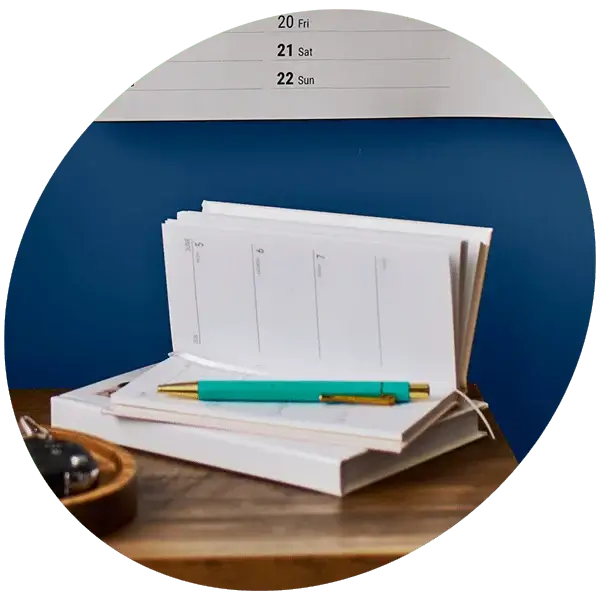 An open diary with a teal pen resting on top, placed on a wooden surface. Behind it is a wall calendar showing dates, and part of a small dish with keys is visible on the side. The background features a blue wall.
