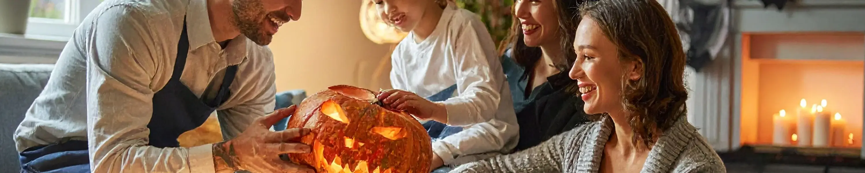 Group of people gathered around a glowing carved pumpkin indoors with candles on a fireplace in the background, creating a warm Halloween atmosphere. Group of people gathered around a glowing carved pumpkin indoors with candles on a fireplace in the background, creating a warm Halloween atmosphere.