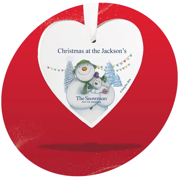 Heart-shaped Christmas ornament with snowman and snowdog illustration, snowy trees, colorful lights, and text Christmas at the Jackson's and The Snowman and The Snowdog. Heart-shaped Christmas ornament with snowman and snowdog illustration, snowy trees, colorful lights, and text Christmas at the Jackson's and The Snowman and The Snowdog.