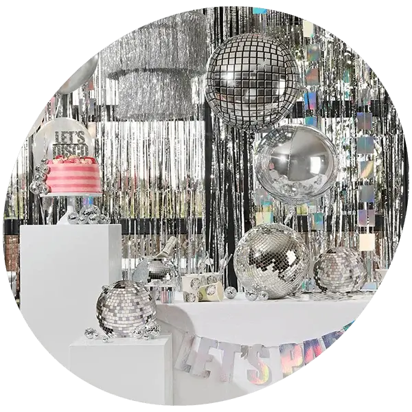 A disco‑themed party setup with mirrored disco balls, silver streamers, metallic decorations and a pink cake displayed on white plinths against a shimmering silver backdrop.