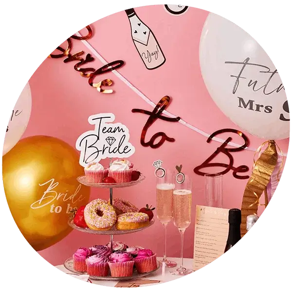 A pink hen party dessert table decorated with themed balloons, a Bride to Be banner, cupcakes, doughnuts, champagne flutes, and party accessories.