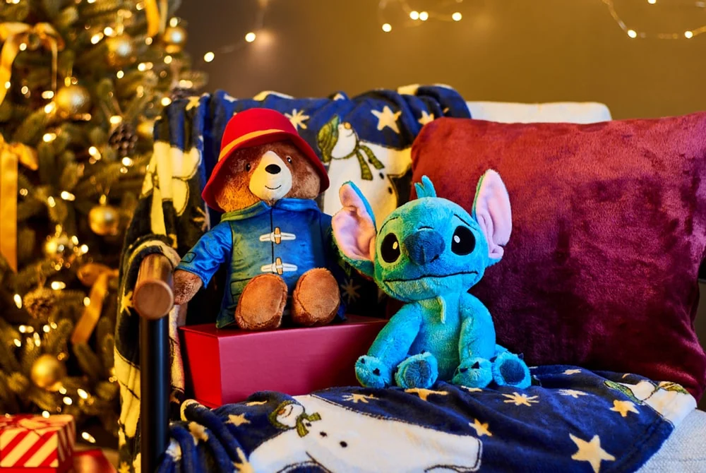 Two plush toys, Paddington Bear in a blue coat and red hat and Stitch in bright blue, sitting on a festive blanket with a Christmas tree and warm lights in the background. Two plush toys, Paddington Bear in a blue coat and red hat and Stitch in bright blue, sitting on a festive blanket with a Christmas tree and warm lights in the background.