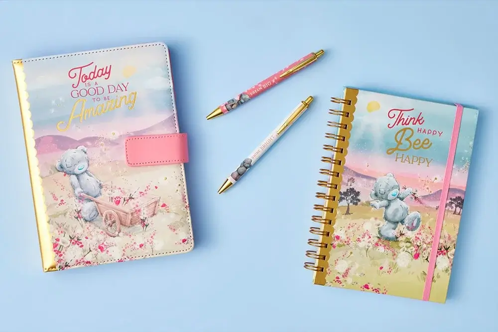 The image shows a pastel-themed stationery set on a light blue background. It includes two notebooks and two pens. The notebook on the left has a cover with an illustration of a bear pushing a cart of flowers, and the text reads 'Today is a good day to be amazing'. The notebook on the right has an illustration of a bear among flowers with mountains in the background, and the text reads 'Think happy bee happy'. Both pens have matching designs featuring bears and floral patterns.