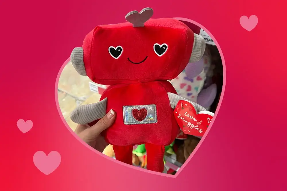 Red plush robot toy with heart‑shaped eyes and a small heart detail on its chest, held in front of a store display.