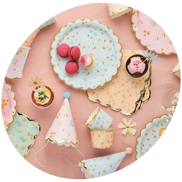 Pastel party tableware set with floral plates, napkins, party hats, cupcakes, and macarons on a pink background