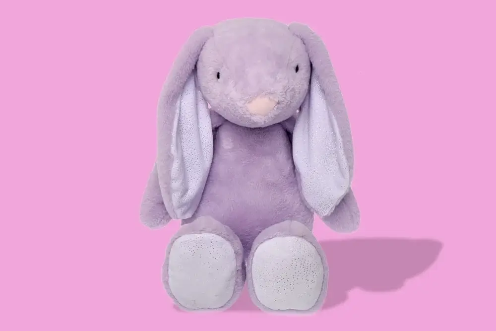 Light purple plush bunny toy with long ears and large feet, sitting against a pink background. Light purple plush bunny toy with long ears and large feet, sitting against a pink background.