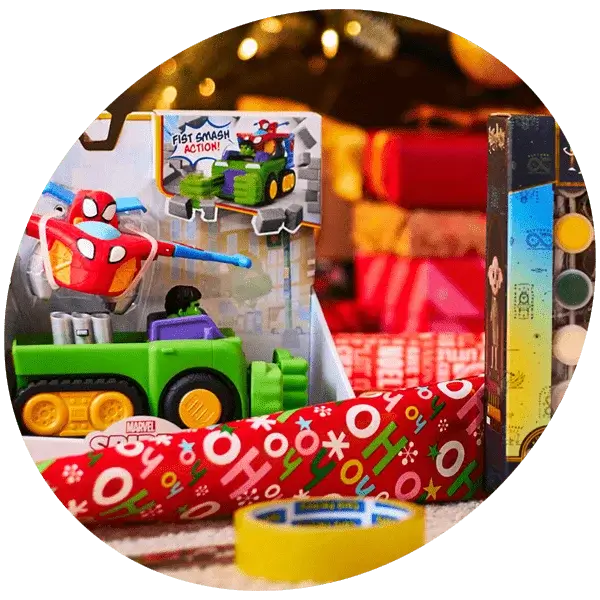 A festive gift-wrapping scene featuring a Marvel toy in its packaging, a roll of red wrapping paper with colorful letters and stars, yellow tape, and other wrapped presents in the background. The setting includes warm holiday lighting and a decorated Christmas tree.