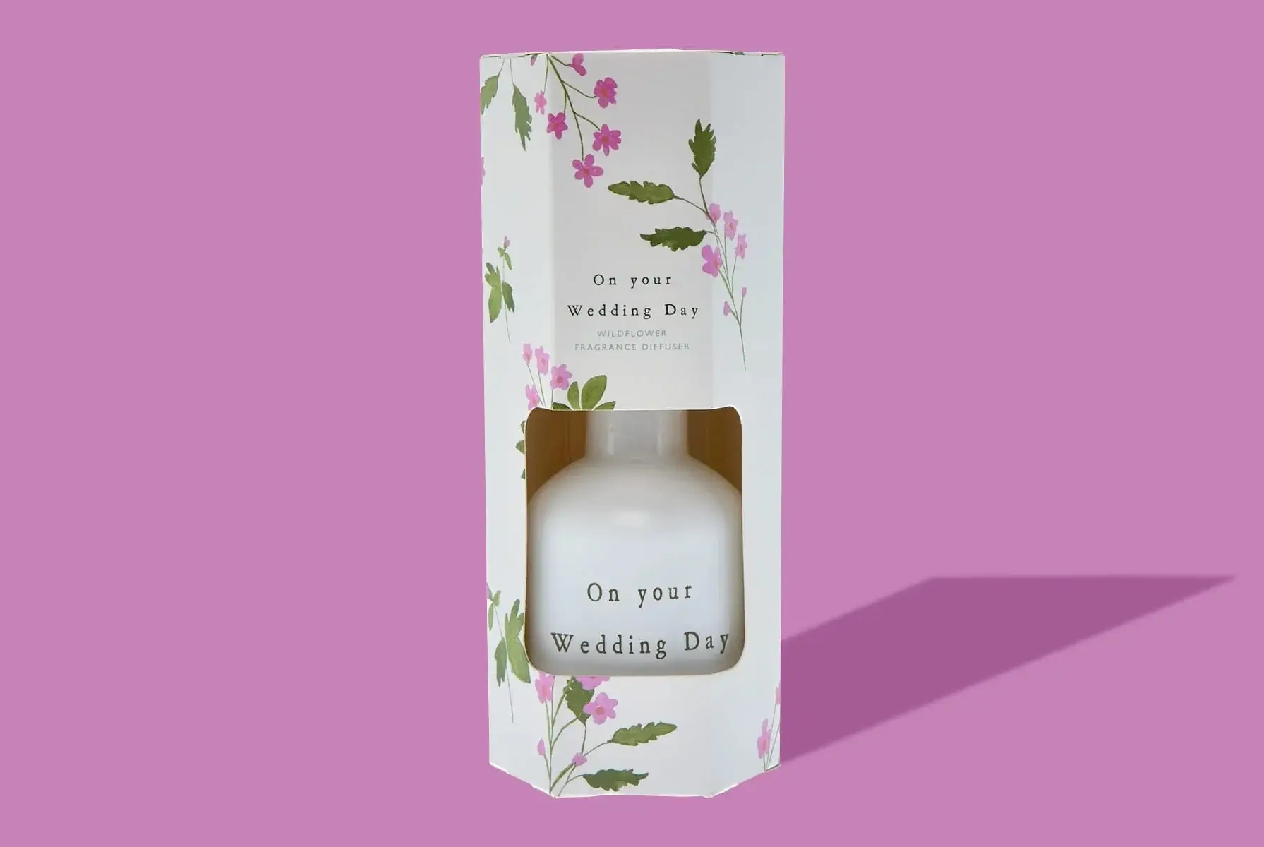 'A white fragrance diffuser bottle with the text 'On your Wedding Day' is partially visible through a rectangular cutout in a floral-patterned box. The box has pink and green floral designs and also displays the text 'On your Wedding Day' along with 'WILDFLOWER FRAGRANCE DIFFUSER'. The background is a solid light purple color.' 'A white fragrance diffuser bottle with the text 'On your Wedding Day' is partially visible through a rectangular cutout in a floral-patterned box. The box has pink and green floral designs and also displays the text 'On your Wedding Day' along with 'WILDFLOWER FRAGRANCE DIFFUSER'. The background is a solid light purple color.'
