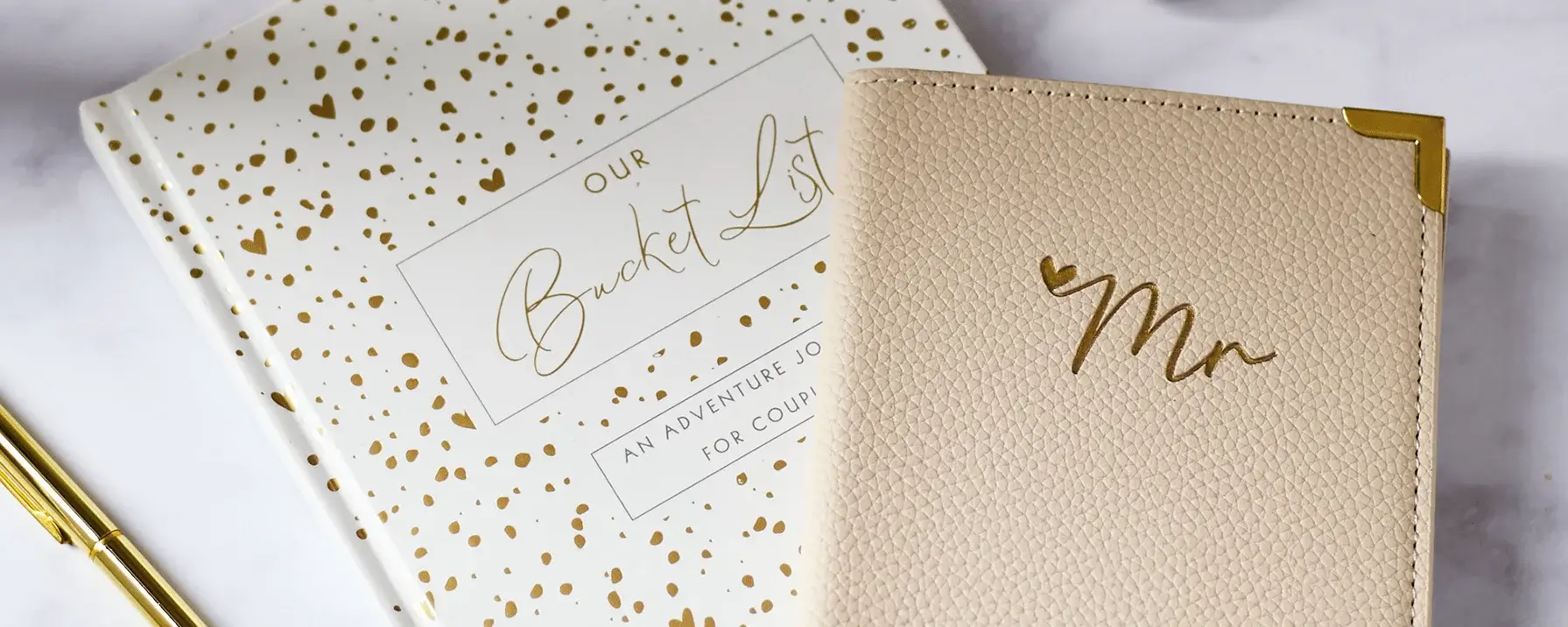 The image shows two notebooks and a pen on a marble surface. One notebook has a white cover with gold polka dots and the text 'OUR Bucket List' and 'AN ADVENTURE JOURNAL FOR COUPLES' written in gold. The other notebook has a beige textured cover with the word 'Mr' written in gold, along with gold corner protectors. A gold pen is placed next to the notebooks. The image shows two notebooks and a pen on a marble surface. One notebook has a white cover with gold polka dots and the text 'OUR Bucket List' and 'AN ADVENTURE JOURNAL FOR COUPLES' written in gold. The other notebook has a beige textured cover with the word 'Mr' written in gold, along with gold corner protectors. A gold pen is placed next to the notebooks.
