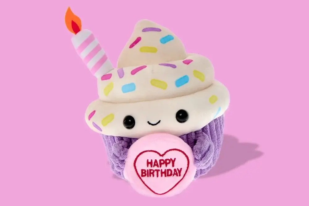 Plush cupcake toy with white frosting, sprinkles, smiling face, and a pink heart that says Happy Birthday, set against a pink background. Plush cupcake toy with white frosting, sprinkles, smiling face, and a pink heart that says Happy Birthday, set against a pink background.