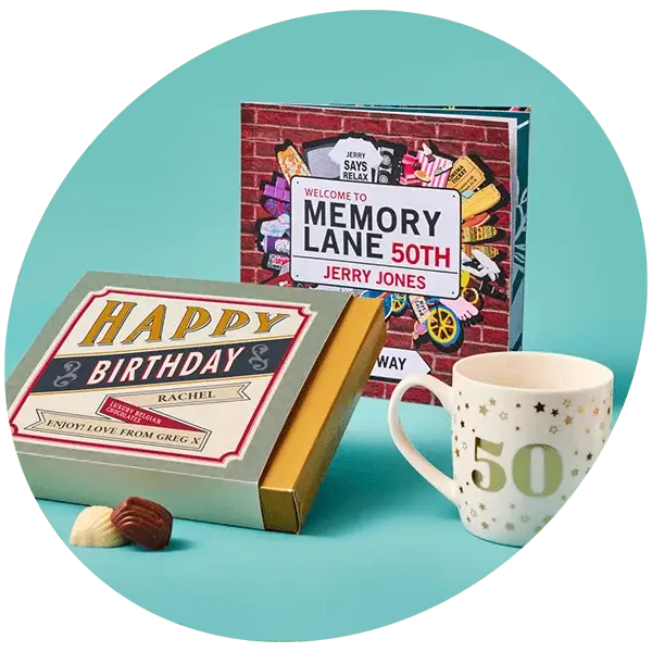 Birthday gift set featuring a book titled 'Welcome to Memory Lane 50th' by Jerry Jones, a white mug with the number '50' in gold, and a box labeled 'Happy Birthday Rachel' with the message 'Enjoy love from Grig x', arranged on a light blue surface with two chocolates beside the box.