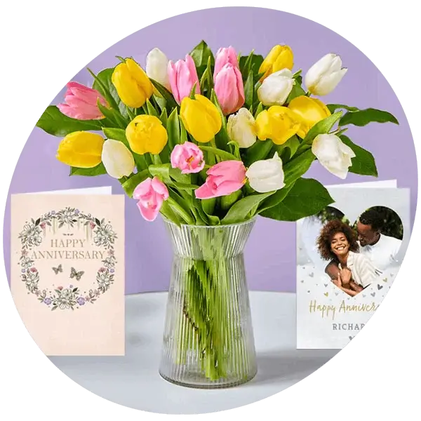Bouquet of pink, yellow, and white tulips in a glass vase with anniversary cards on a purple background