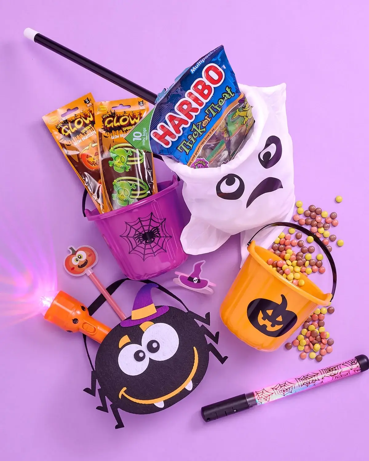 Halloween-themed items on a purple background including candy-filled buckets with spider and pumpkin designs, a ghost container, a flashlight, and scattered sweets. Halloween-themed items on a purple background including candy-filled buckets with spider and pumpkin designs, a ghost container, a flashlight, and scattered sweets.