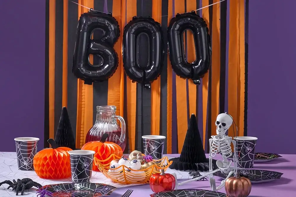Halloween table setting with black and orange decorations, including BOO balloon letters, spider web cups and plates, a red drink pitcher, pumpkins, a skeleton figure, and a candy basket. Halloween table setting with black and orange decorations, including BOO balloon letters, spider web cups and plates, a red drink pitcher, pumpkins, a skeleton figure, and a candy basket.