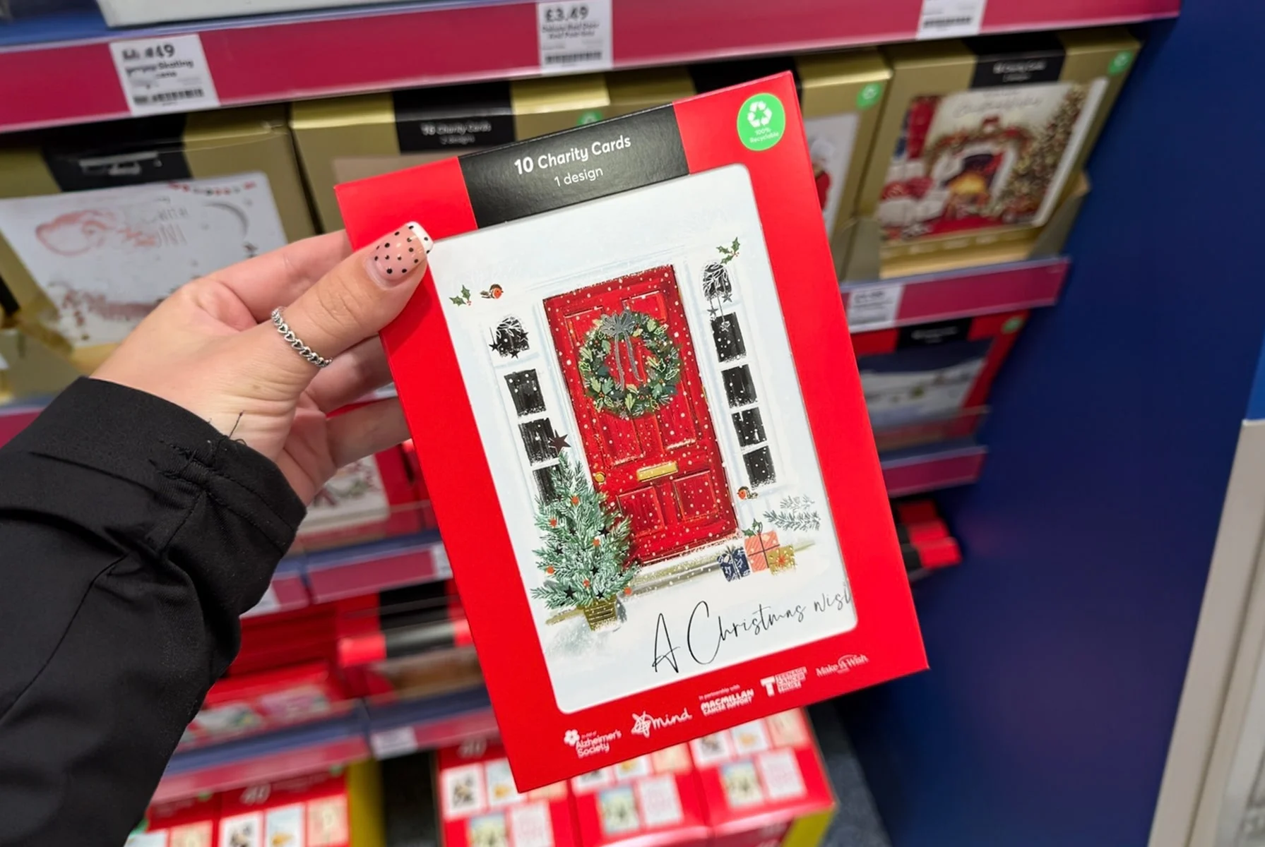 A hand holding a pack of charity Christmas cards featuring a red door design with a wreath and festive decorations, displayed in a store aisle. A hand holding a pack of charity Christmas cards featuring a red door design with a wreath and festive decorations, displayed in a store aisle.