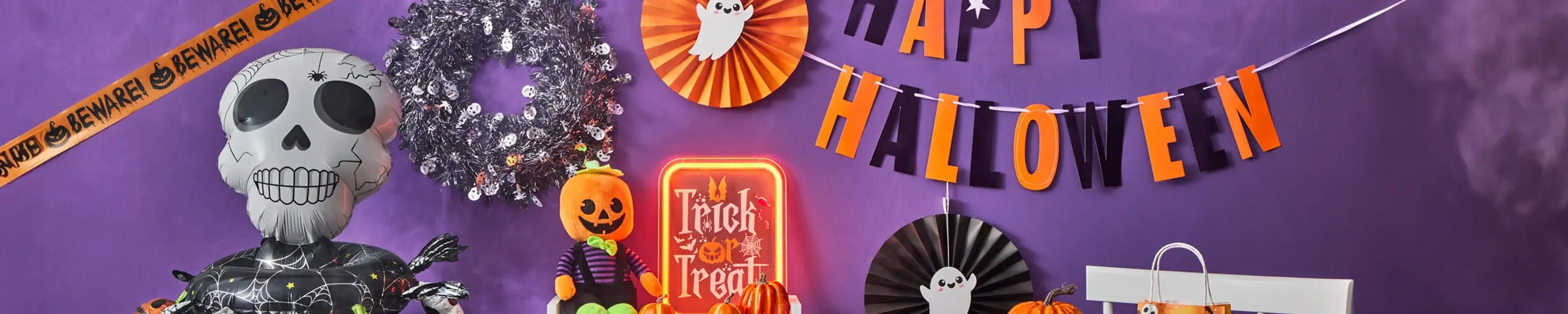 Halloween display with an inflatable skeleton, caution tape, silver wreath, pumpkins, Trick or Treat sign, and Happy Halloween banner with ghost cutouts on a purple background.