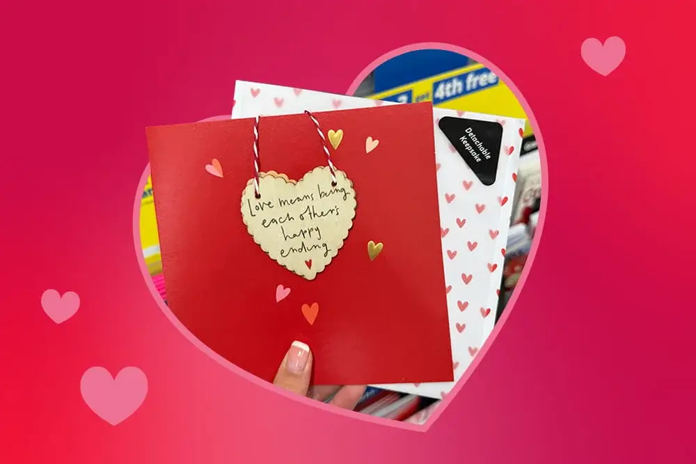 Red Valentine’s card with a heart‑shaped tag that reads love means being each other’s happy ending, held in front of a display of other heart‑patterned cards.