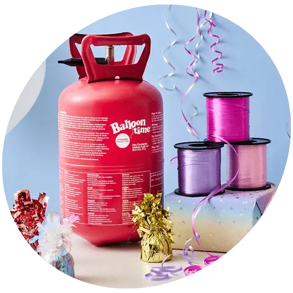 A red helium tank labeled 'Balloon Time' is placed on a table. Surrounding the tank are colorful party decorations, including three spools of ribbon in pink, purple, and lavender, metallic gift bows in gold and silver, and curling ribbons hanging down from above.