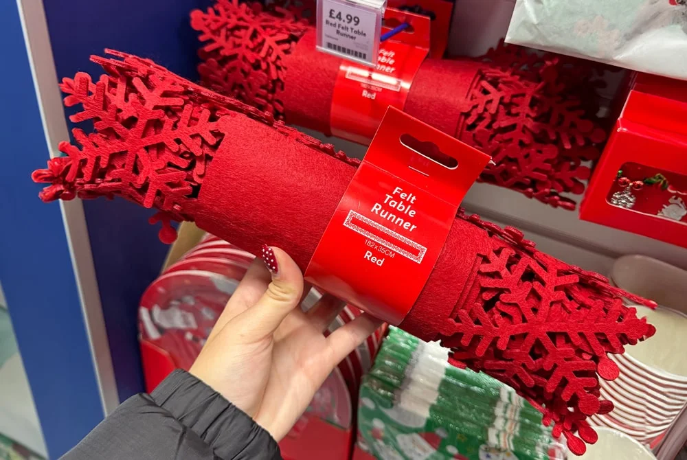 A hand holding a rolled red felt table runner with decorative snowflake cutouts, packaged with a label reading Felt Table Runner, displayed among other festive tableware in a store. A hand holding a rolled red felt table runner with decorative snowflake cutouts, packaged with a label reading Felt Table Runner, displayed among other festive tableware in a store.