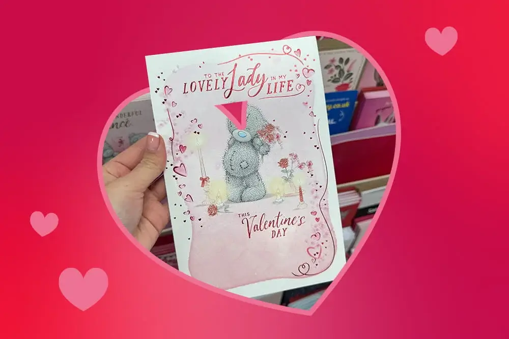 Valentine’s card featuring a cute teddy bear surrounded by pink hearts and floral decorations, held in front of a card display.