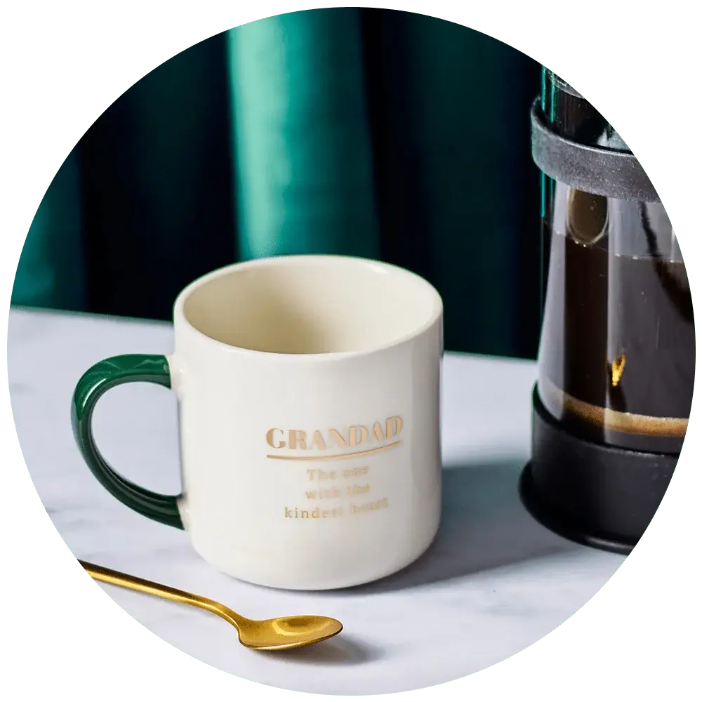 A white mug with a green handle is placed on a marble surface. The mug has the word 'GRANDAD' written in gold, followed by the phrase 'The one with the kindest heart'. Next to the mug is a gold spoon and part of a French press coffee maker filled with coffee. The background features teal-colored curtains.' A white mug with a green handle is placed on a marble surface. The mug has the word 'GRANDAD' written in gold, followed by the phrase 'The one with the kindest heart'. Next to the mug is a gold spoon and part of a French press coffee maker filled with coffee. The background features teal-colored curtains.'