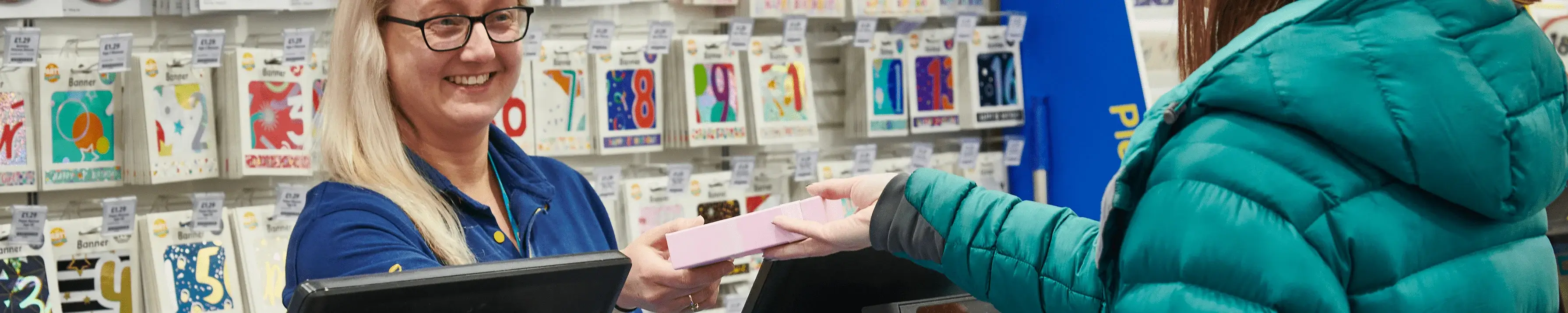 A person in a green jacket hands a pink box to a cashier wearing a blue uniform. The background shows shelves filled with various greeting cards.' A person in a green jacket hands a pink box to a cashier wearing a blue uniform. The background shows shelves filled with various greeting cards.'