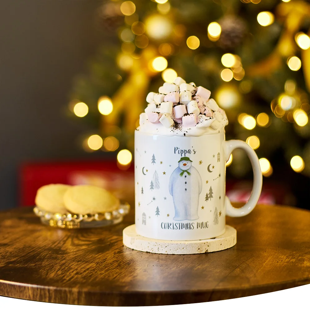 Christmas mug with marshmallows and whipped cream on a wooden table, featuring a snowman and the text 'Pippa's Christmas Mug', with a blurred tree and festive lights in the background.