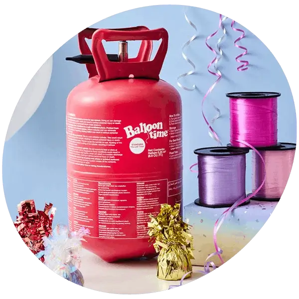 A red helium tank labeled 'Balloon Time' is placed on a table. Surrounding the tank are colorful party decorations, including three spools of ribbon in pink, purple, and lavender, metallic gift bows in gold and silver, and curling ribbons hanging down from above.
