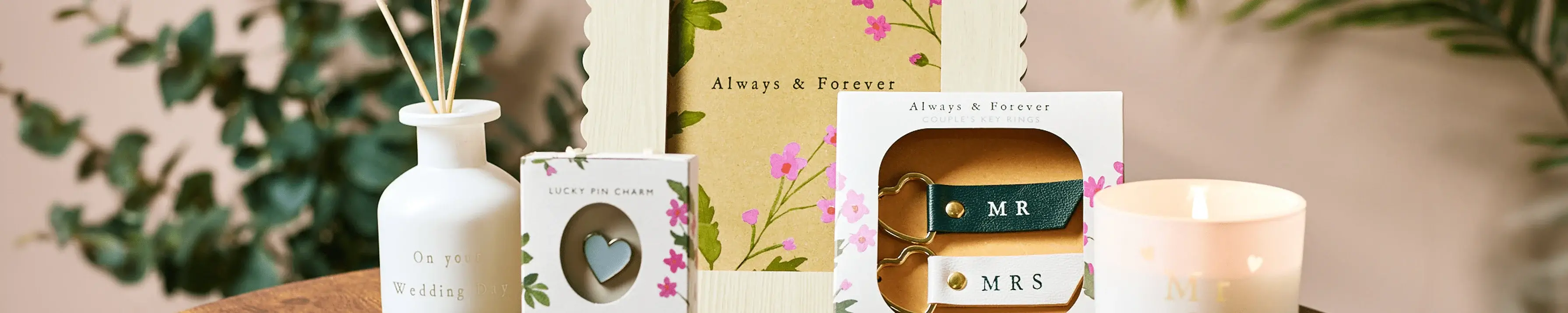 The image shows a collection of wedding-themed items arranged on a table. There is a small white vase with reed diffusers, a box labeled 'Our Wedding' with floral designs, a greeting card with the text 'Always & Forever', and two keychains in boxes labeled 'MR' and 'MRS'. A lit candle is also present on the right side of the arrangement. The image shows a collection of wedding-themed items arranged on a table. There is a small white vase with reed diffusers, a box labeled 'Our Wedding' with floral designs, a greeting card with the text 'Always & Forever', and two keychains in boxes labeled 'MR' and 'MRS'. A lit candle is also present on the right side of the arrangement.