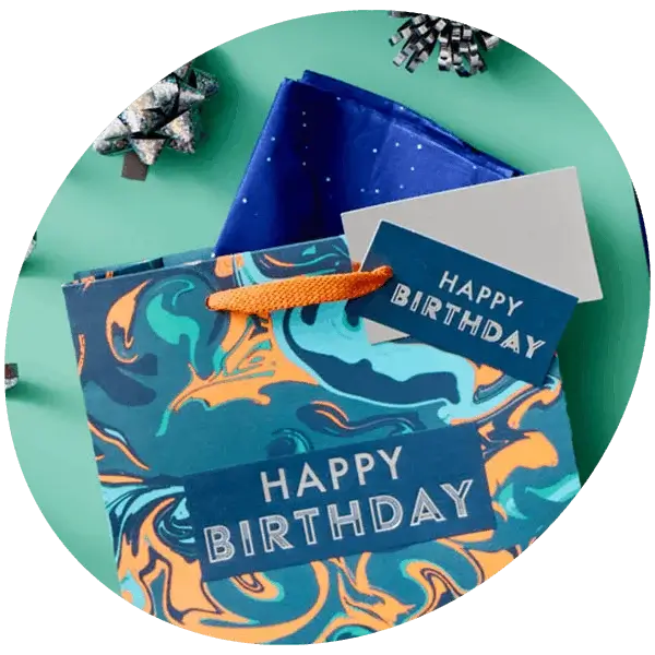 A colourful birthday gift bag with a marbled blue and orange design, blue tissue paper and a matching Happy Birthday tag, surrounded by silver bows on a green background.