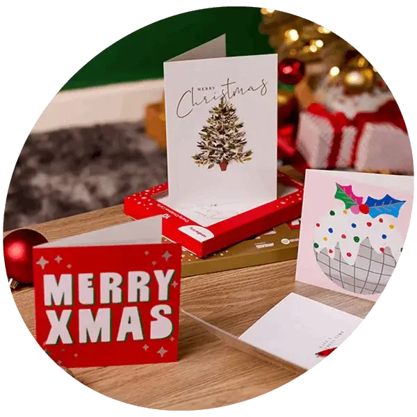 Selection of Christmas greeting cards displayed on a wooden table with festive decorations, wrapped gifts, and a decorated Christmas tree in the background.