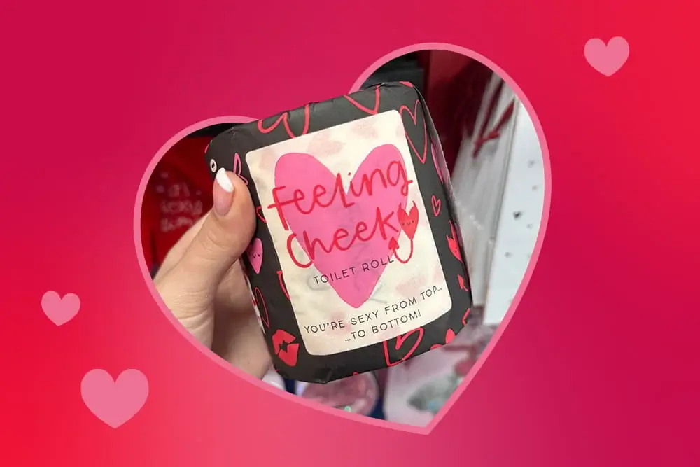Valentine‑themed toilet roll wrapped in black and pink packaging with a large heart and the phrase feeling cheeky, held up in front of store shelves.