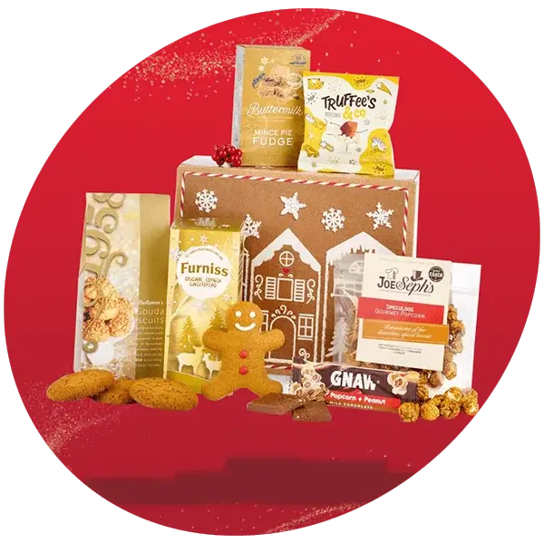 Festive gift hamper displayed against a red background, featuring a gingerbread-themed box surrounded by assorted treats including biscuits, mince pie fudge, truffles, gourmet popcorn, and a gingerbread cookie.