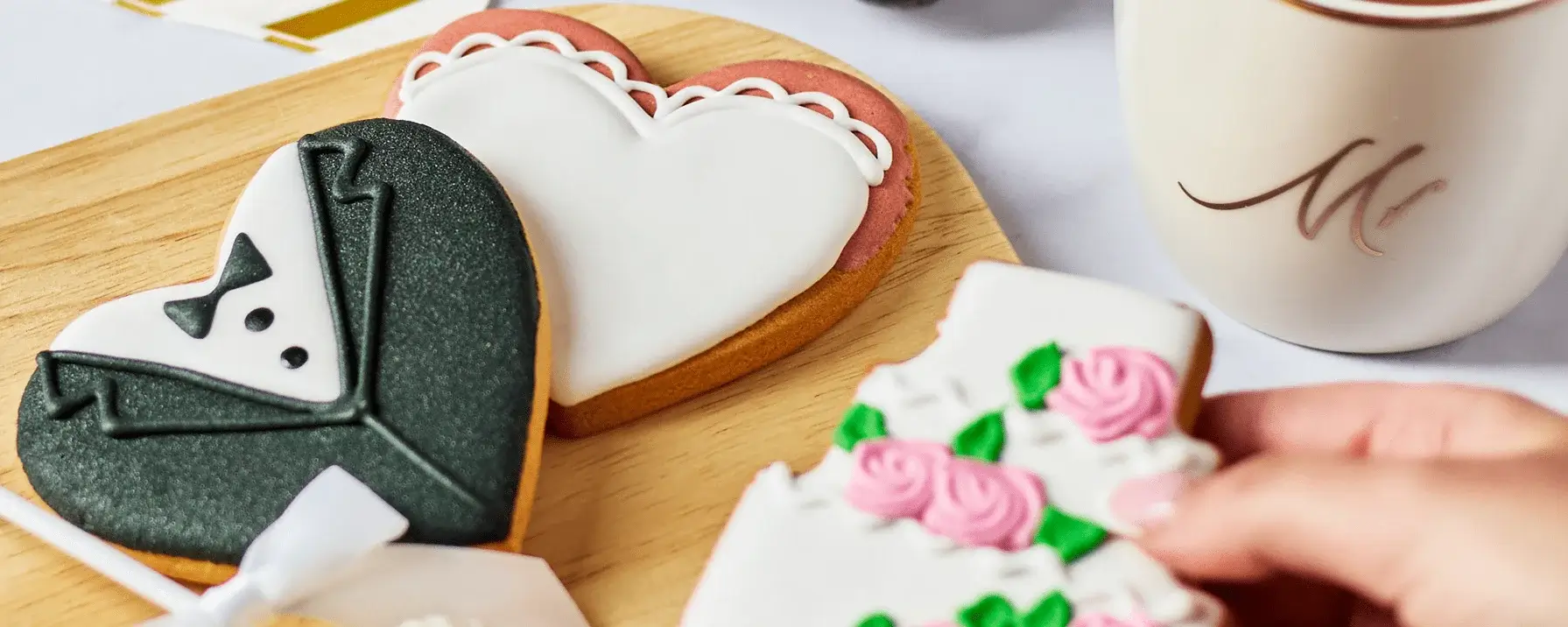 A close-up image of two heart-shaped cookies on a wooden tray. One cookie is decorated to look like a tuxedo, and the other is decorated with white icing and pink roses. A hand is holding the cookie with pink roses, which has a bite taken out of it. There is also a cup with the letter 'M' on it in the background. A close-up image of two heart-shaped cookies on a wooden tray. One cookie is decorated to look like a tuxedo, and the other is decorated with white icing and pink roses. A hand is holding the cookie with pink roses, which has a bite taken out of it. There is also a cup with the letter 'M' on it in the background.
