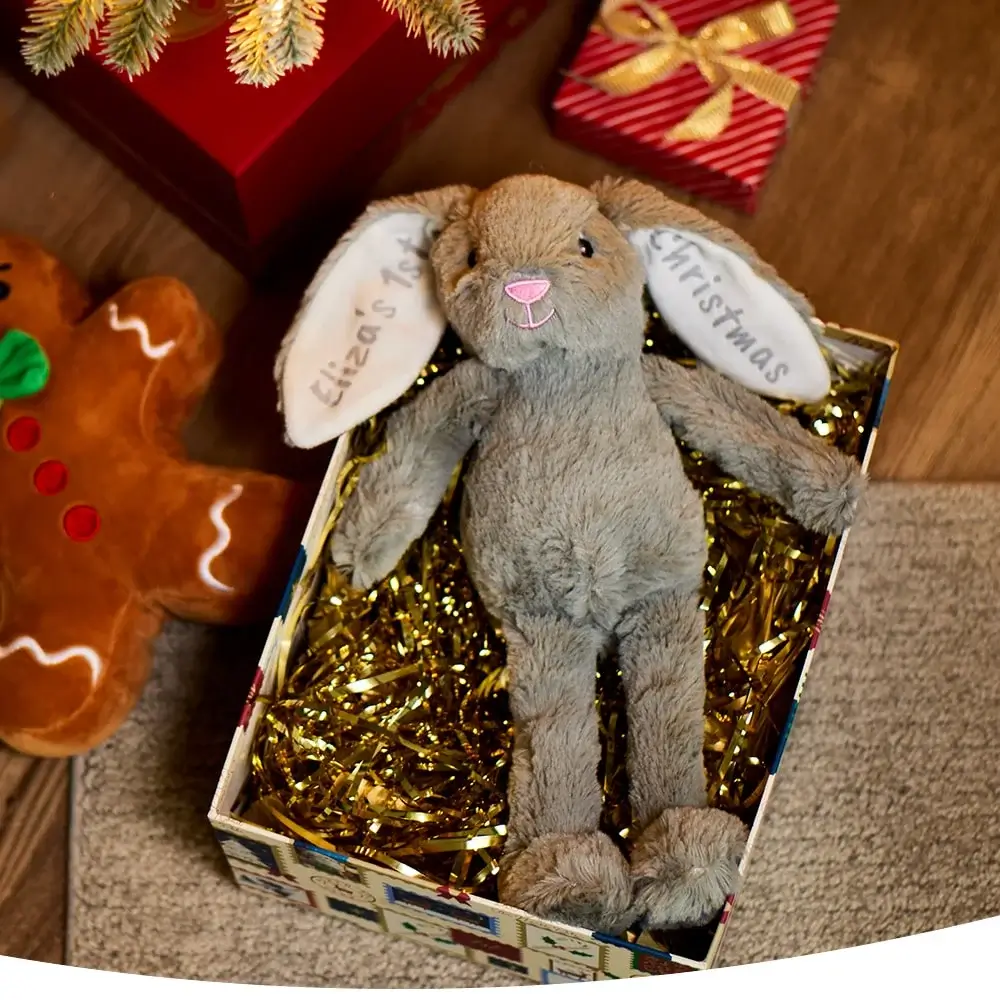 Plush bunny toy with 'Eliza's 1st Christmas' on its ears in a festive gift box, surrounded by gold tinsel, a gingerbread plush, wrapped presents, and a decorated Christmas tree.