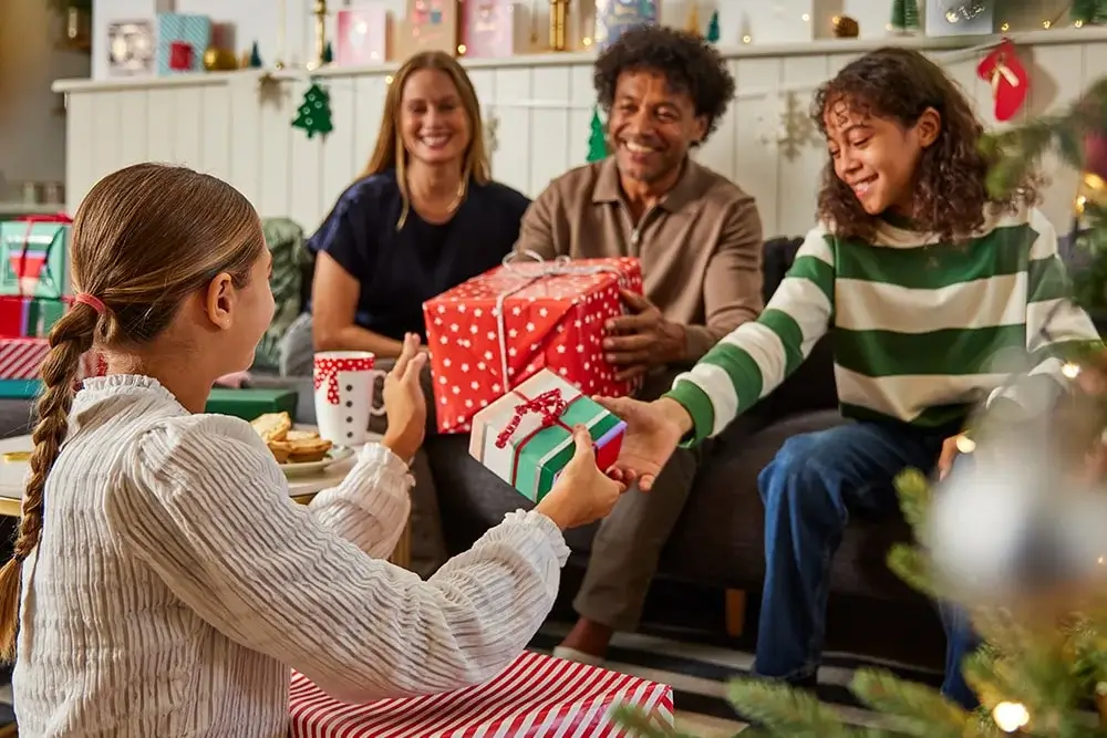 A festive living room scene with a decorated Christmas tree in the foreground and several wrapped presents on the floor. People are sitting together exchanging gifts, including a large red box with white snowflake patterns. The background features holiday decorations on the wall. A festive living room scene with a decorated Christmas tree in the foreground and several wrapped presents on the floor. People are sitting together exchanging gifts, including a large red box with white snowflake patterns. The background features holiday decorations on the wall.