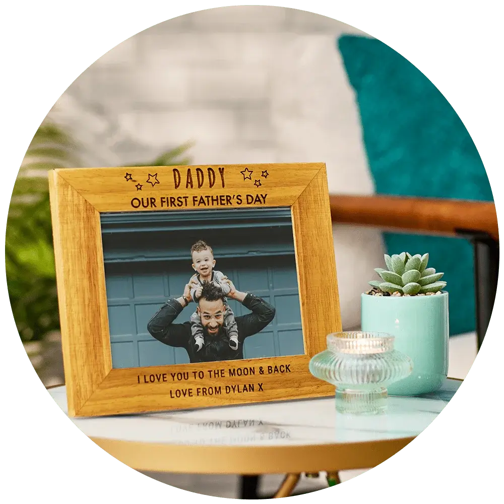 A wooden picture frame with the text 'DADDY' at the top, 'OUR FIRST FATHER'S DAY' in the middle, and 'I LOVE YOU TO THE MOON & BACK LOVE FROM DYLAN X' at the bottom. The frame contains a photo of an adult holding a child on their shoulders. The frame is placed on a table next to a small potted succulent plant and a glass candle holder.' A wooden picture frame with the text 'DADDY' at the top, 'OUR FIRST FATHER'S DAY' in the middle, and 'I LOVE YOU TO THE MOON & BACK LOVE FROM DYLAN X' at the bottom. The frame contains a photo of an adult holding a child on their shoulders. The frame is placed on a table next to a small potted succulent plant and a glass candle holder.'