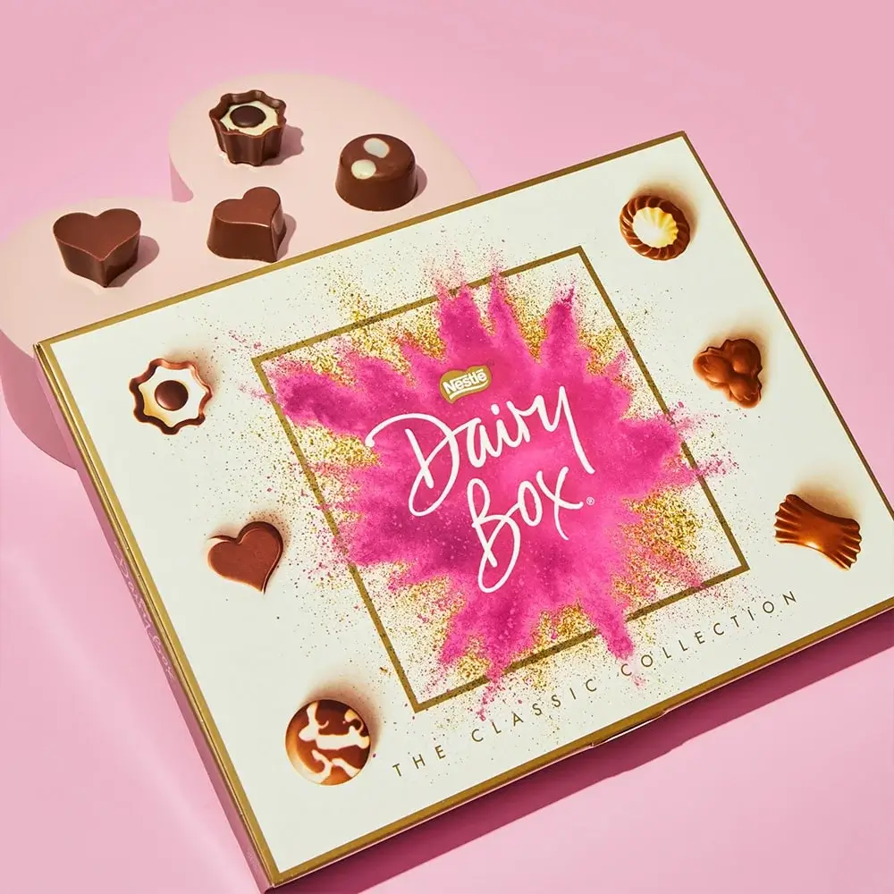 Nestlé Dairy Box The Classic Collection chocolate box with a pink and gold splash design on the lid, surrounded by assorted chocolates in various shapes, displayed on a pink background with a heart-shaped platform.