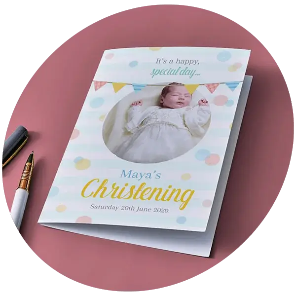 A christening card featuring pastel polka dots, a photo window showing a baby in a white gown, and text reading Maya’s Christening alongside the date, displayed on a pink surface with pens nearby.