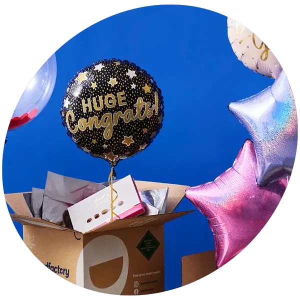 An open cardboard box filled with tissue paper and a small gift box, surrounded by colorful balloons. The central balloon is black with gold stars and the text 'HUGE Congrats!'
