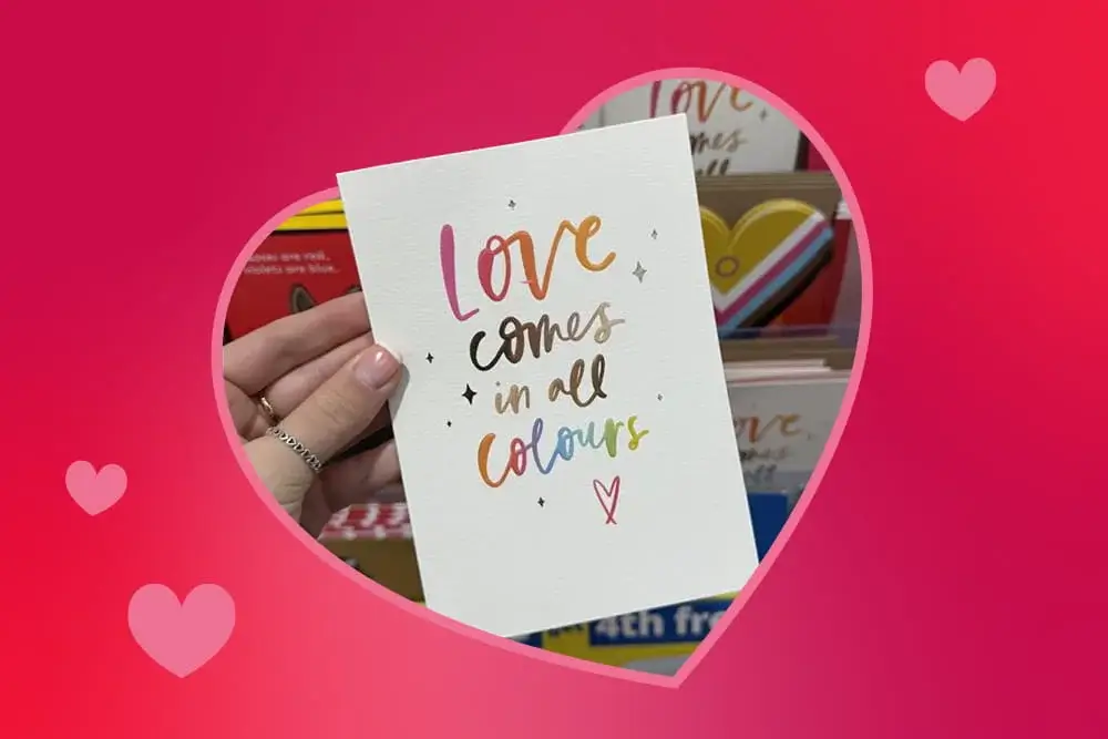 Valentine’s card with rainbow lettering that says love comes in all colours, held in front of a display of other colourful cards.
