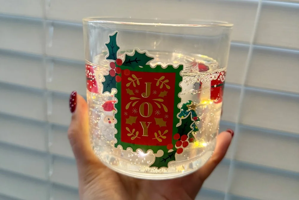 A clear glass decorated with holly leaves, berries, and the word JOY in festive colors, held in front of window blinds with light reflecting through the water inside. A clear glass decorated with holly leaves, berries, and the word JOY in festive colors, held in front of window blinds with light reflecting through the water inside.