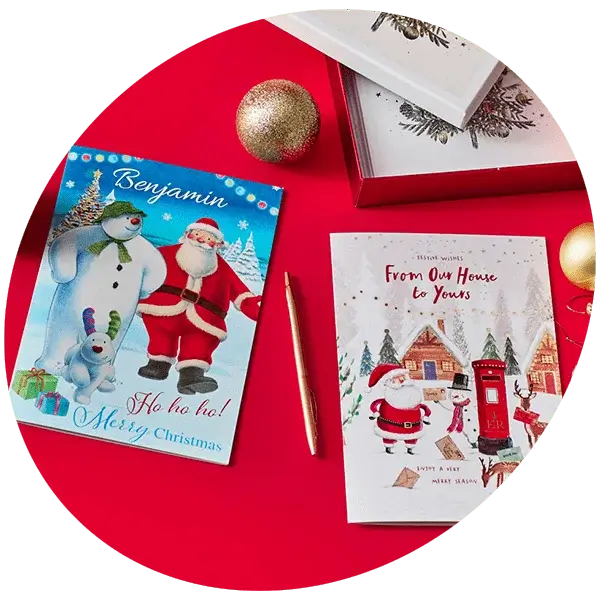 Two Christmas cards on a red surface with a gold pen, festive ornaments, and a gift box in the background.