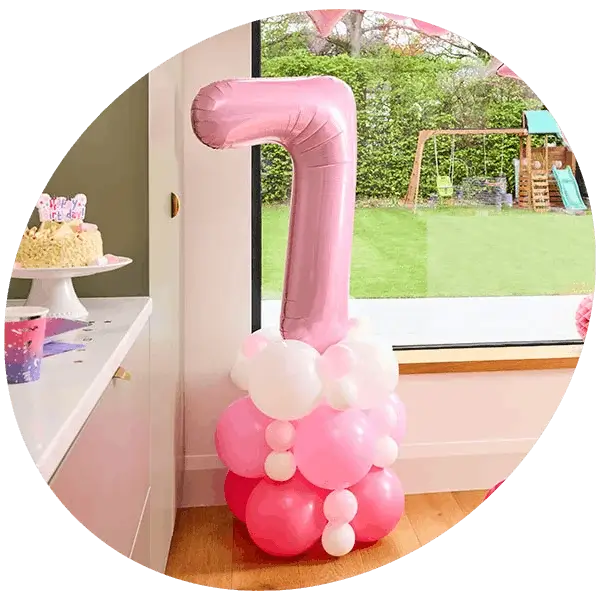 Pink number 7 foil balloon display with a matching balloon base, positioned beside a birthday cake and party decorations.
