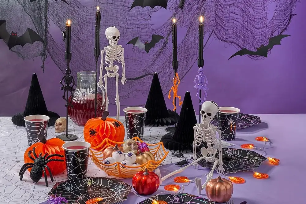 Halloween table setup with skeletons, black witch hats, orange pumpkins, spider webs, and candles on a purple background. Halloween table setup with skeletons, black witch hats, orange pumpkins, spider webs, and candles on a purple background.