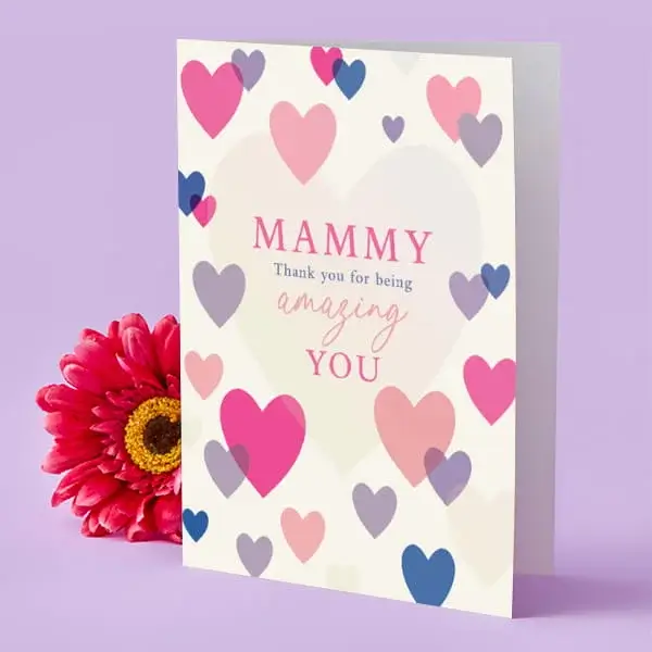 A cream Mother’s Day card decorated with pink, purple, and blue hearts, displayed upright beside a bright pink flower.