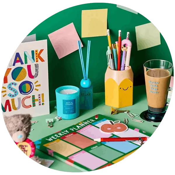 Colourful desk scene with stationery, weekly planner, pens in a holder, sticky notes, candle, diffuser, and a glass on a green background